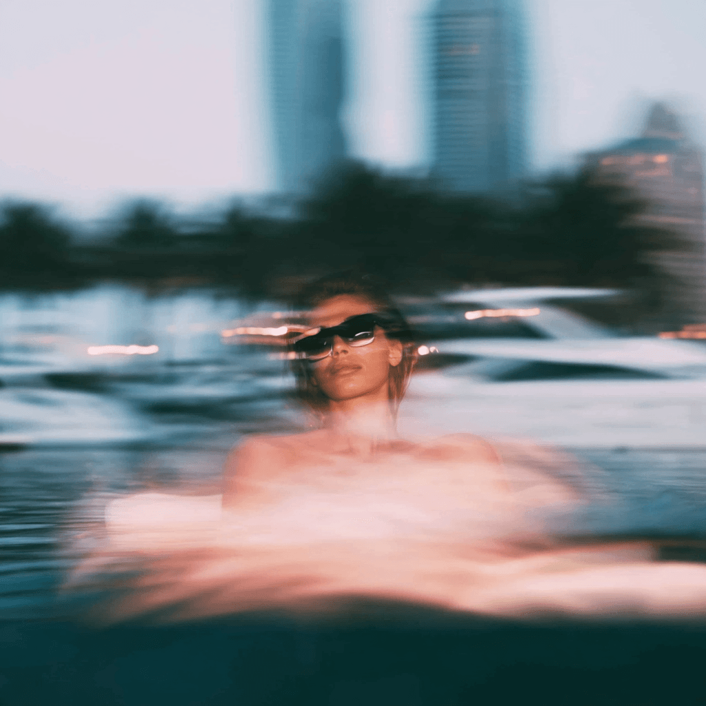 Motion-blurred figure in sunglasses in the water at dusk, with yachts and a city skyline behind.