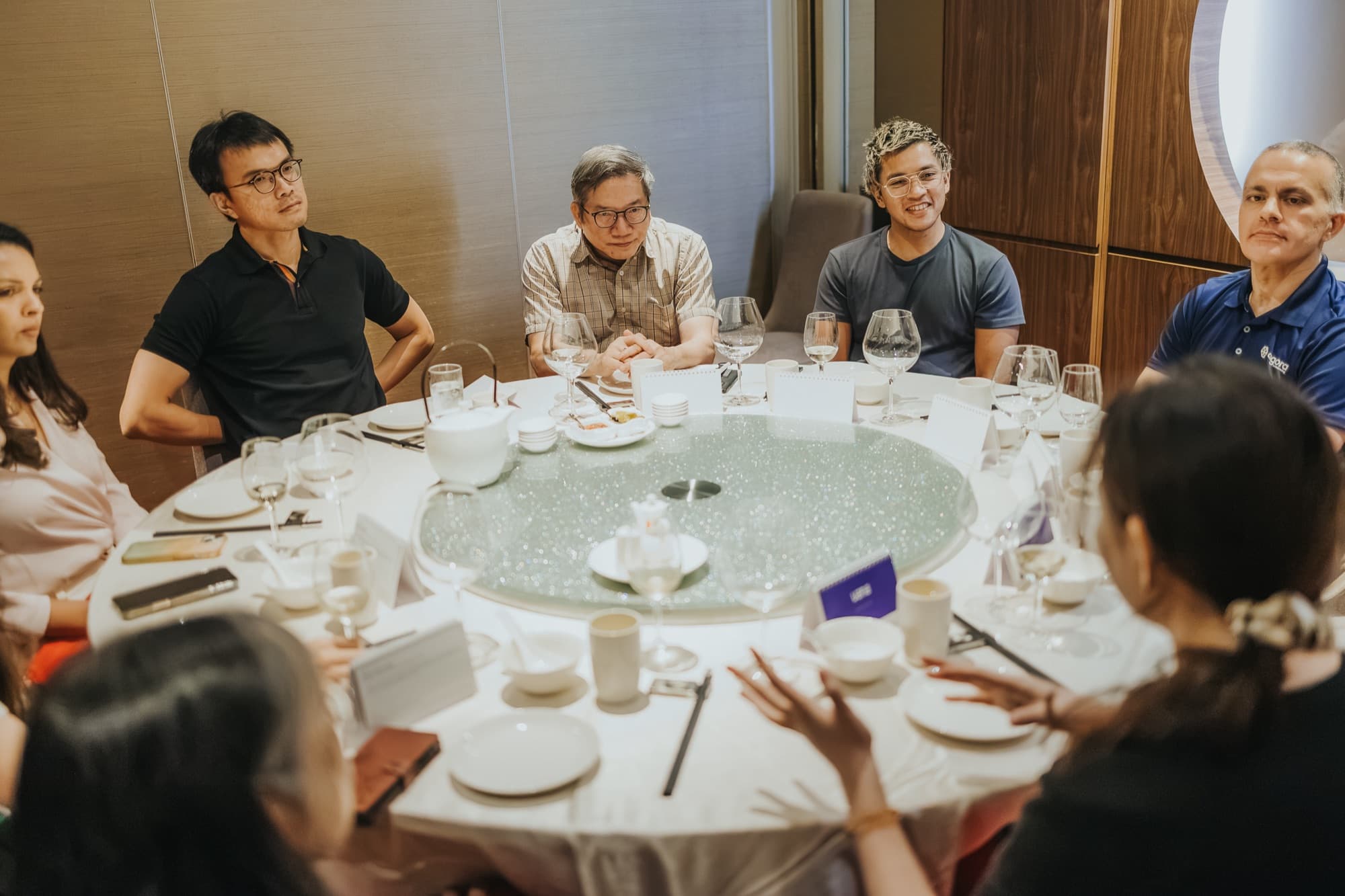 Roundtable participants seated around a large round dining table set with glasses and tea, listening to one person speak.