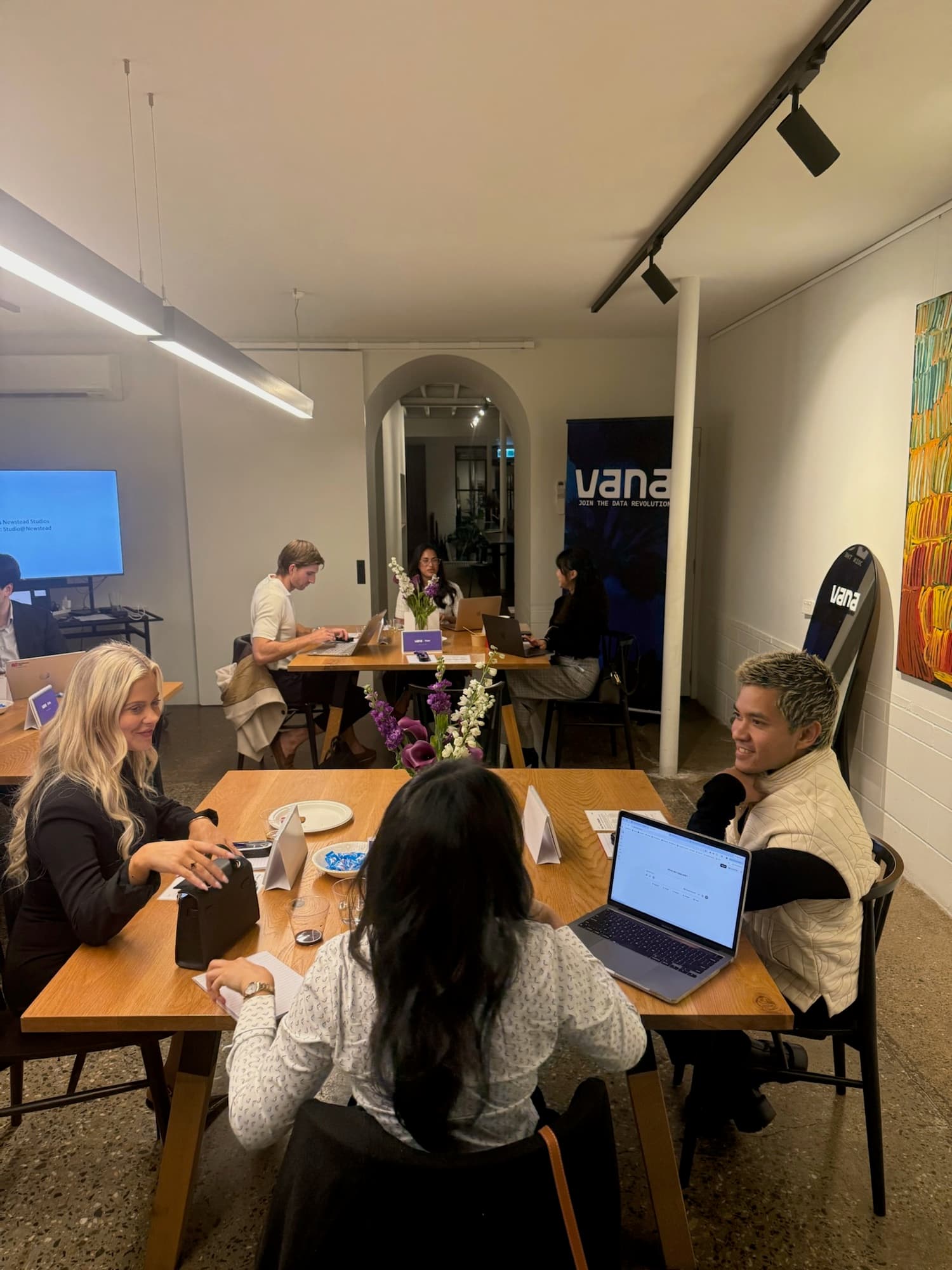 Mixed teams working on laptops at shared tables, with a Vana banner in the background.