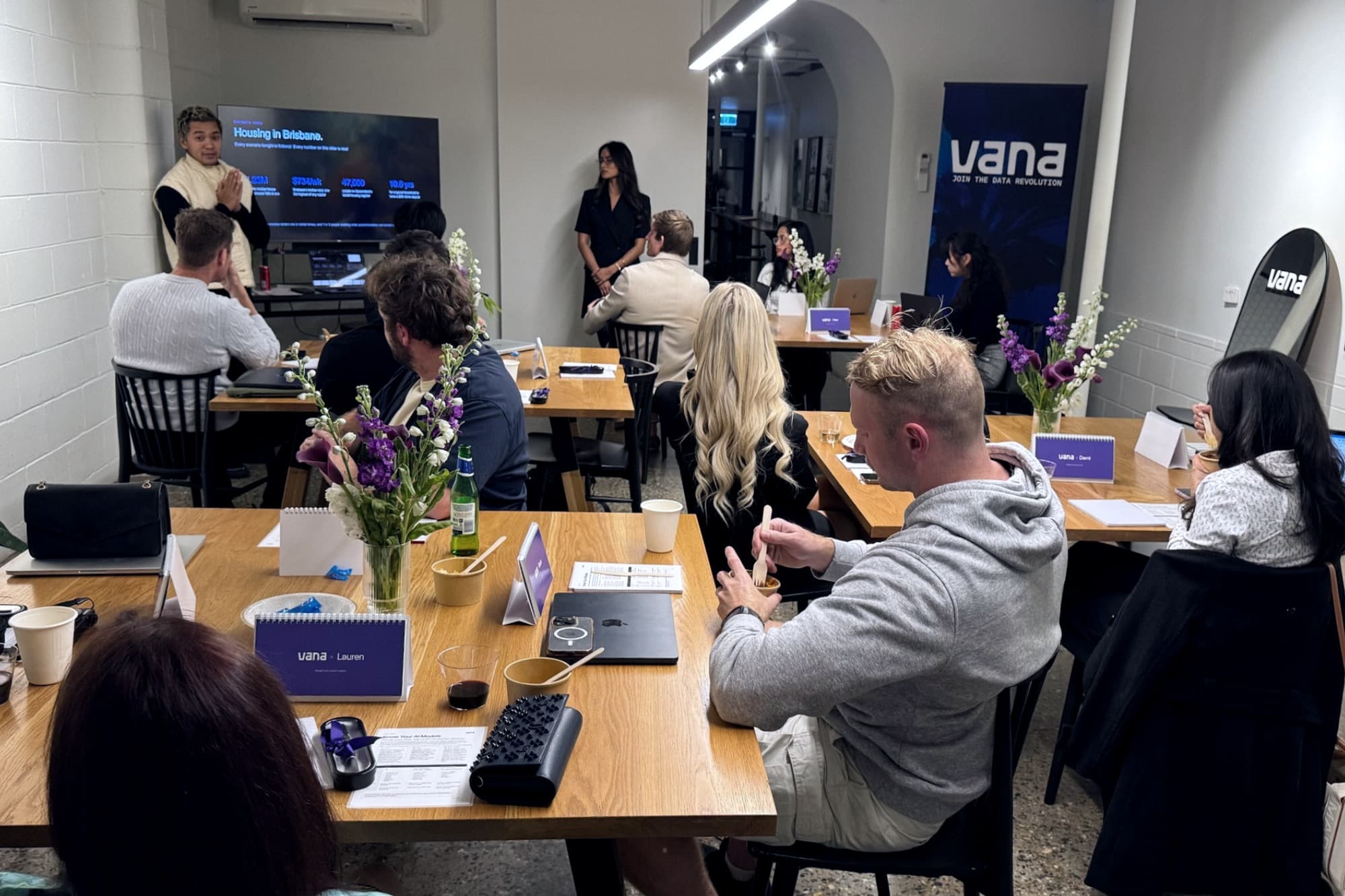 Participants seated at shared tables in a Brisbane workshop, facing a presenter and a slide headed 'Housing in Brisbane', with Vana branding on a banner behind them.